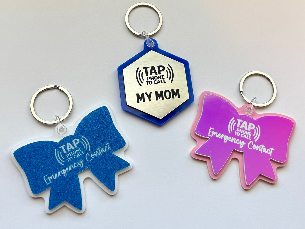 Three tap tag keychains with blue bow, silver hexagon, and pink bow on a light gray background
