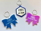 Three tap tag keychains with blue bow, silver hexagon, and pink bow on a light gray background